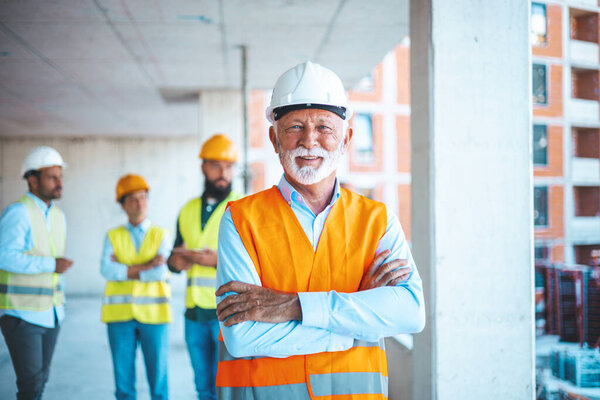 Portrait of a construction site worker in hard hat and safety vest. Senior architect at a construction site smiling at camera - Incidental people working at background. 