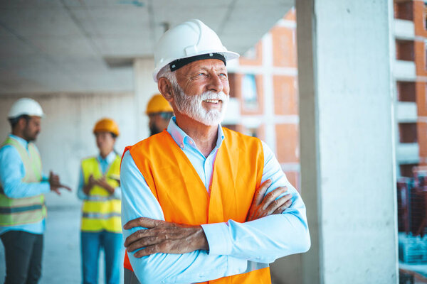 Visiting construction site. Portrait of confident elderly construction engineer in classic suit and hardhat is holding digital tablet and looking at camera while visiting building object