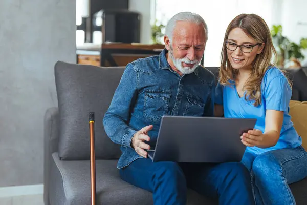 Nurse help senior man with internet and laptop on the sofa in a retirement home. Healthcare worker, caregiver or medical professional helping retired man with email and technology in the lounge