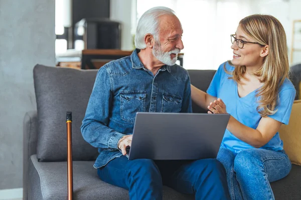 Senior man with disability learning to pay bills online with laptop together with social worker. Nurse help senior retirement man working with laptop. Active senior man using laptop with female carer sitting by on sofa at home