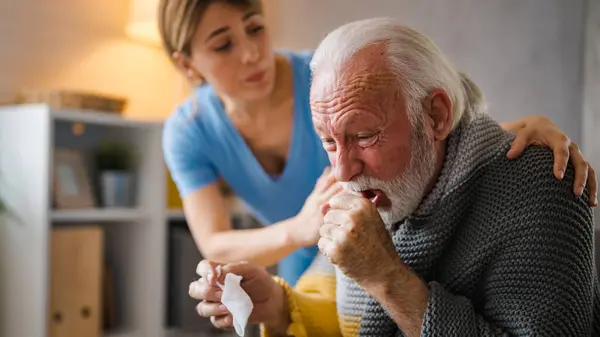 Cold exhausted senior man with flu wrapped in a warm blanket blowing his nose with a tissue in the livingroom. Nurse take care of him. Geriatrician helping lonely elderly sick man.