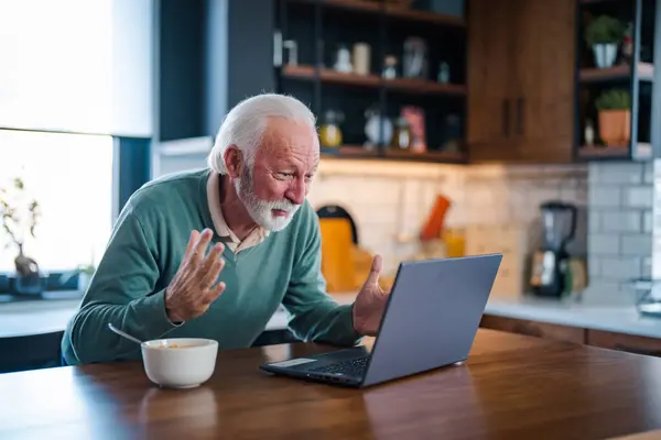 Good-looking enterpreneur working on laptop while having coffee and breakfast. Elderly man using laptop at kitchen table. Computer, money and communication with an elderly male pensioner in the kitchen