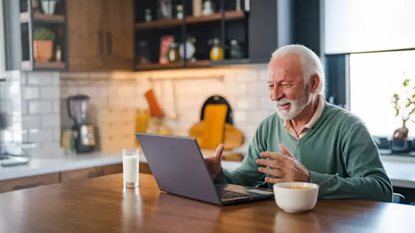 Cheerful senior man during video conference in kitchen on the laptop while enjoying breakfast and a cup of coffee. Elderly person using internet online chat technology video webcam making a video call