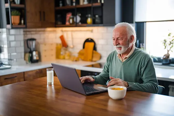 Close up of a senior man using a laptop in the kitchen. Shot of a senior man sitting alone and using his laptop to calculate his finances at home. Portrait of senior man using laptop at home