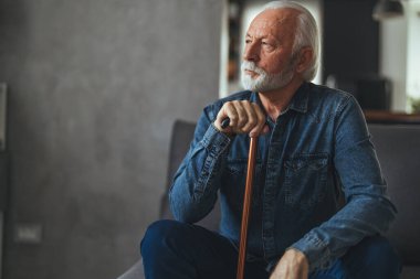 Engelli sopalı gri saçlı adam kanepede oturuyor ve evde düşünüyor.