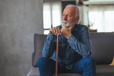 Engelli sopalı gri saçlı adam kanepede oturuyor ve evde düşünüyor.