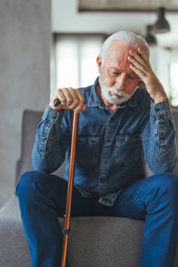 Engelli sopalı gri saçlı adam kanepede oturuyor ve evde düşünüyor.