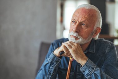 Engelli sopalı gri saçlı adam kanepede oturuyor ve evde düşünüyor.