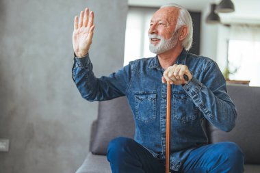 Engelli çubuğu olan olgun bir adam kanepede oturuyor ve evde el sallıyor.