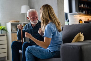 Genç bayan hemşire ve evdeki kanepede sakat sopayla oturan yaşlı adam.