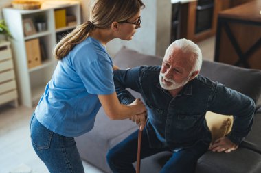 Hemşire engelli ihtiyara yardım ediyor. Evdeki kanepeden kalkmış sopa yiyor.