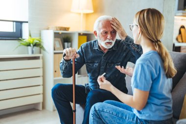Termometresi olan genç bayan hemşire evdeki kanepede sakat çubuklu yaşlı adamla konuşuyor.