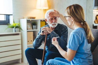 Genç bayan hemşire evdeki kanepede engelli çubuğu olan yaşlı bir adamın vücut ısısını ölçüyor.