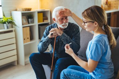 Genç bayan hemşire evdeki kanepede engelli çubuğu olan yaşlı bir adamın vücut ısısını ölçüyor.