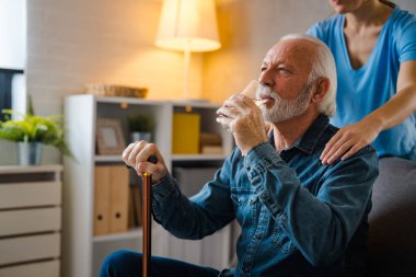 Kısmi hemşire engelli yaşlı bir adamın omuzlarında oturuyor. Evdeki kanepede camdan su içiyor.