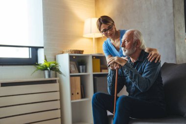 Gülümseyen genç bayan hemşire evdeki kanepede sakat sopalı olgun bir adama bakıyor.
