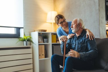 Gülümseyen genç bayan hemşire evdeki kanepede sakat sopalı olgun bir adama bakıyor.