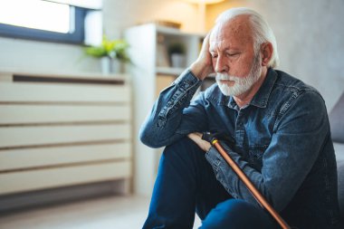 Üzüntülü kıdemli adam engelli sopasıyla evdeki kanepenin yanında oturuyor.