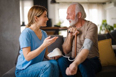 Genç bayan hemşire evdeki kanepede engelli sopalı yaşlı adamın baskısını ölçüyor.