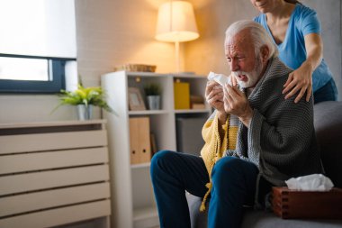 Hastalığın omuzlarında el ele tutuşan kısmi hemşire. Evdeki kanepede peçeteye hapşıran yaşlı adam.