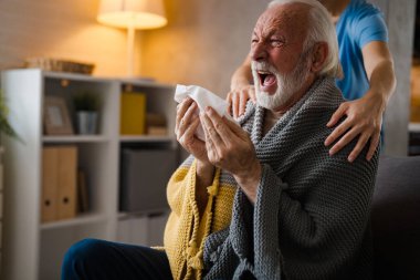 Hastalığın omuzlarında el ele tutuşan kısmi hemşire. Evdeki kanepede peçeteye hapşıran yaşlı adam.