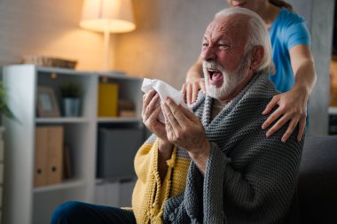 Hastalığın omuzlarında el ele tutuşan kısmi hemşire. Evdeki kanepede peçeteye hapşıran yaşlı adam.