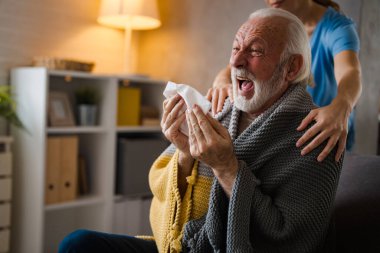 Hastalığın omuzlarında el ele tutuşan kısmi hemşire. Evdeki kanepede peçeteye hapşıran yaşlı adam.