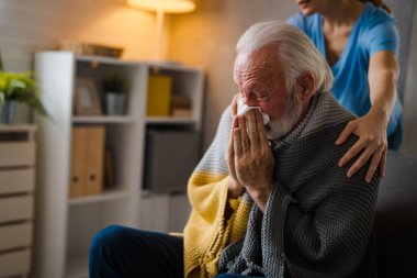 Hastalığın omuzlarında el ele tutuşan kısmi hemşire. Evdeki kanepede peçeteye hapşıran yaşlı adam.
