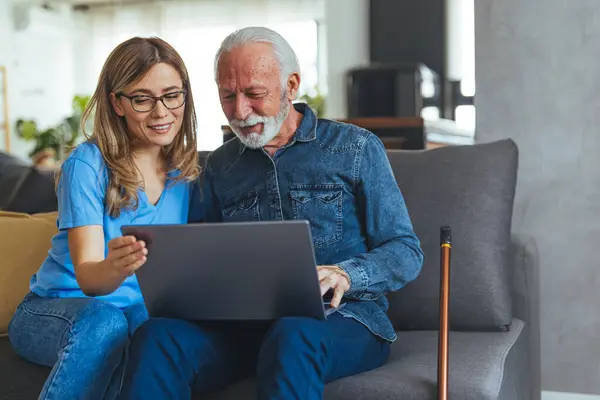Active senior man using laptop with female carer sitting by on sofa at home. Nurse help senior man with internet and laptop on the sofa in a retirement home. Healthcare worker, caregiver or medical professional helping