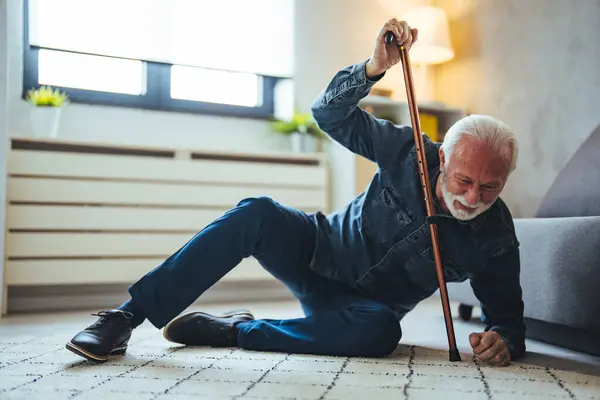 Elder senior man lying on floor after falling down with wooden walking stick beside couch on rug in living room at home. Old man suffering with pain and struggling to get up after falling down at home
