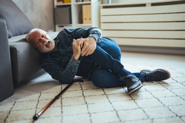 Injured senior male groaning in pain while he tries to get up with a stick after fall at home. He holds his lower back and can barely stand up. Senior Man Fallen On Carpet With Walking Stick