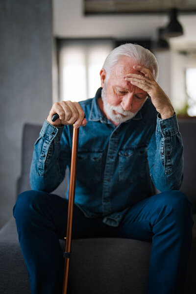 Седой волосатый мужчина с инвалидной палкой сидит на диване и думает дома