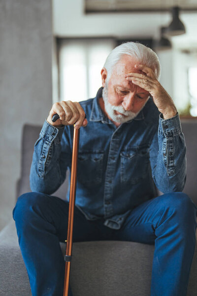 Седой волосатый мужчина с инвалидной палкой сидит на диване и думает дома