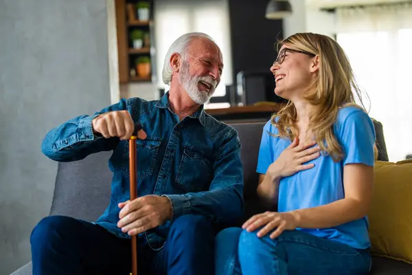 Young female nurse and senior man with disabled stick sitting and laughing on sofa at home