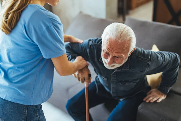 Nurse helping senior man with disabled stick standing up from sofa at home