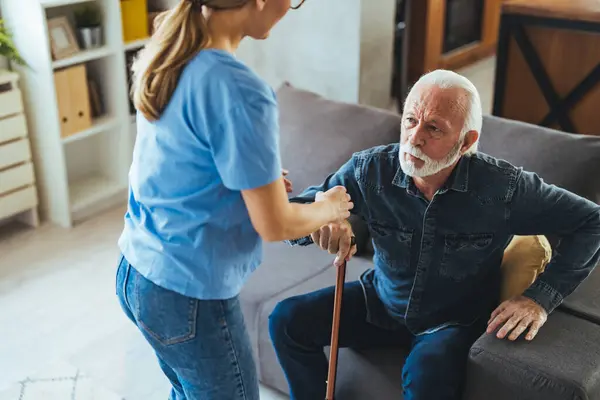 Nurse helping senior man with disabled stick standing up from sofa at home