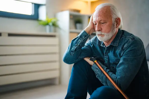 Sadness senior man with disabled stick sitting on floor near sofa at home