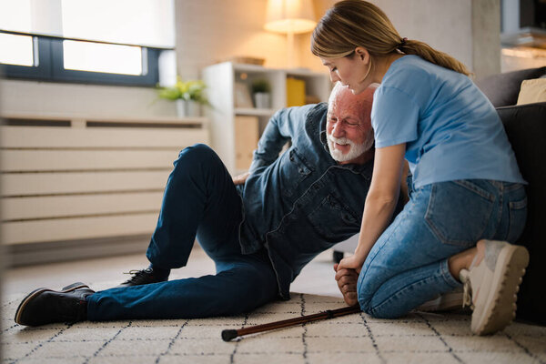 Young female nurse helping senior man standing up from floor at home