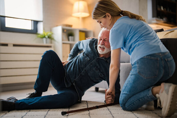 Young female nurse helping senior man standing up from floor at home