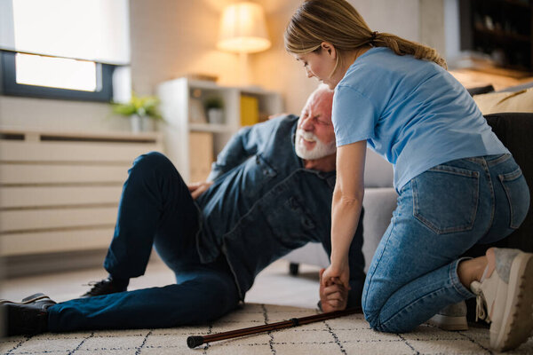 Young female nurse helping senior man standing up from floor at home