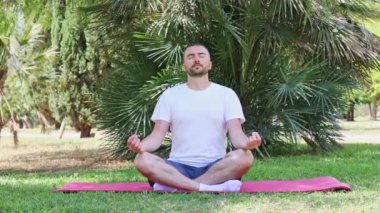 Orta yaşlı atletik bir adam, şehir parkında bir minder üzerinde lotus pozisyonunda otururken yoga yapıyor. Erkek, gözleri kapalı meditasyon yapıyor. Spor salonu açık havada..