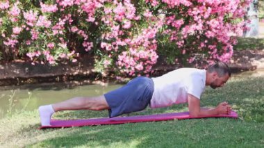 Şehir parkındaki bir paspasın üzerinde tahta egzersizi yapan orta yaşlı bir atletik. Spor salonu açık havada..