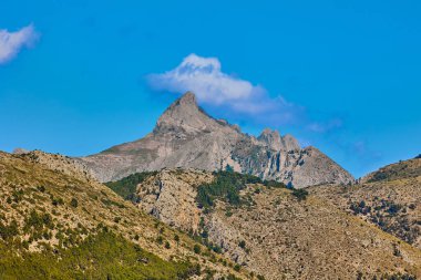 Mavi gökyüzü ve küçük bulutlarla güneşli bir günde kayalık bir dağın manzarası.