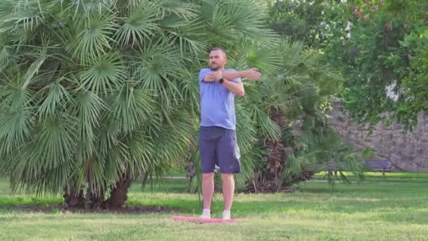 中年の白人男性は屋外の公園でフィットネスマットでストレッチ演習を行います 健康的なアクティブライフスタイル — ストック動画