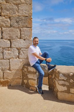Kafkasyalı bir adam Akdeniz kıyısındaki bir ortaçağ şatosunun taş duvarına yaslanmış oturuyor. Valencia, Peniscola. İspanya.