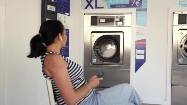 洗濯物が終わるのを待っている洗濯物に座っている若い女性の後ろの景色 — ストック動画