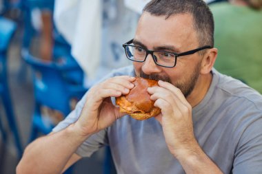 Gözlüklü genç bir adam küçük bir burger yer.