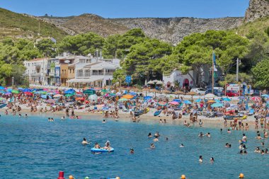 Granadella plajı, Javea, Alicante, İspanya - 12 Ağustos 2023: Sıcak bir günde çam ağaçlarının olduğu bir körfezde küçük bir çakıllı plaj, yüksek turizm sezonunda insanlarla doludur..