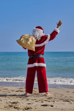 Sıcak bir yaz gününde Noel Baba denizde. Noel Baba 'nın denizin önündeki kumlu bir sahilde çıplak ayakla dikiz aynasından görünüşü. Omzunda hediyelerle bir çanta tutuyor ve elini sallıyor..