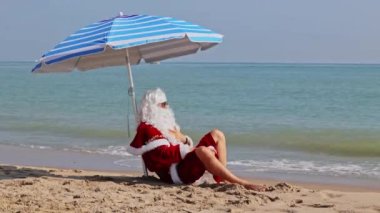 Noel Baba bir plaj şemsiyesinin altında denizin arka planında oturur ve sıcak bir yaz gününün tadını çıkarır. Sıcak ülkelerde Noel ya da yeni yıl tatili. Noel Baba 'nın yaz tatili.